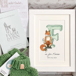 Baby gift box with green watercolour alphabet letter in white frame and green knitted baby booties. Booklet included