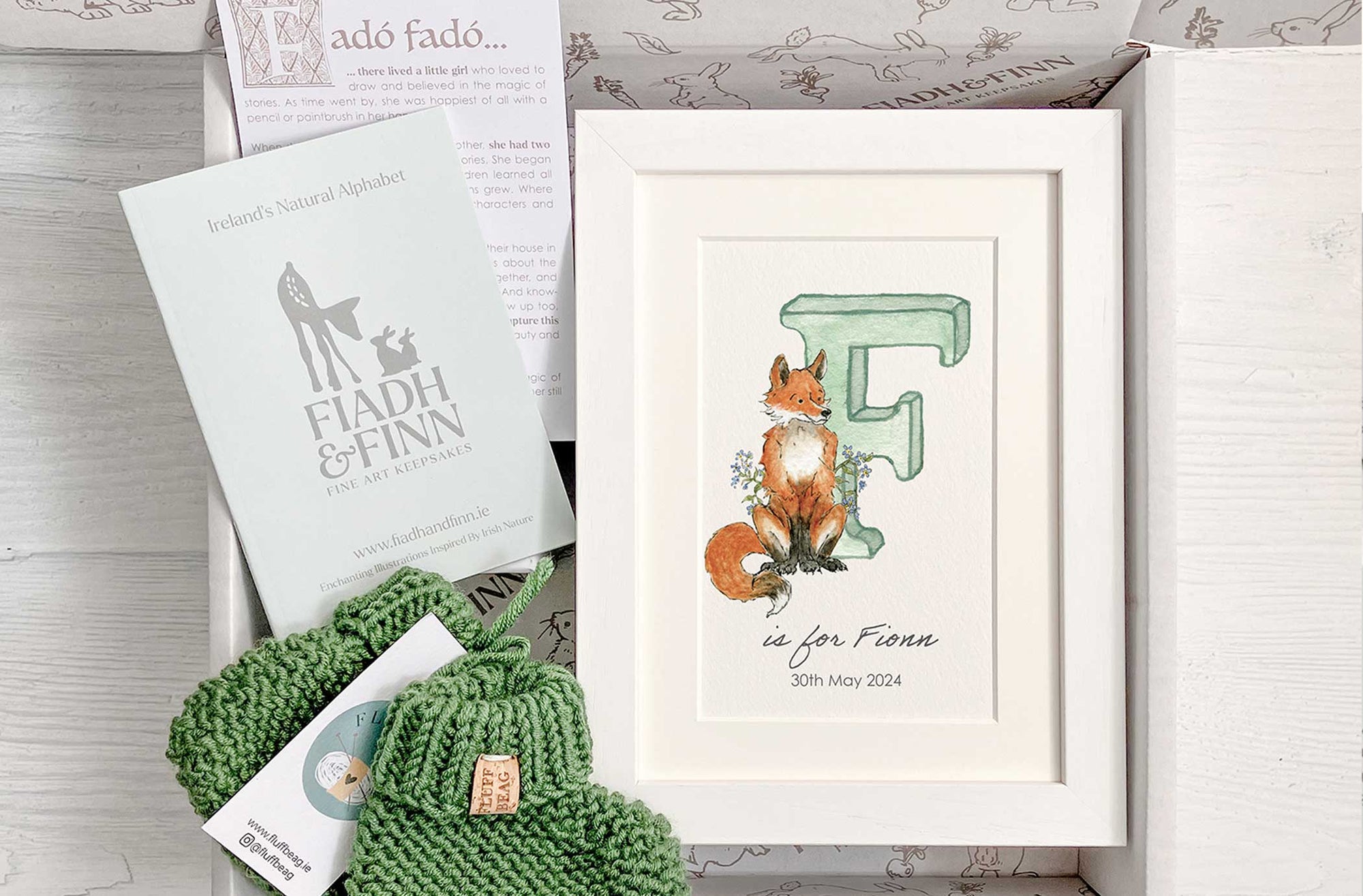 Baby gift box with green watercolour alphabet letter in white frame and green knitted baby booties. Booklet included