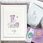 Baby gift box with violet watercolour alphabet letter in white frame and violet knitted baby booties. Booklet included